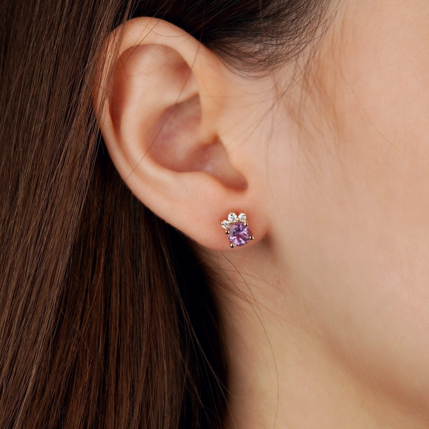 A close-up of a person's ear wearing a small stud earring with a purple heart-shaped gemstone and a silver-toned floral setting-delicate earrings-feminine jewelry- gold jewelry gift- amethyst jewelry