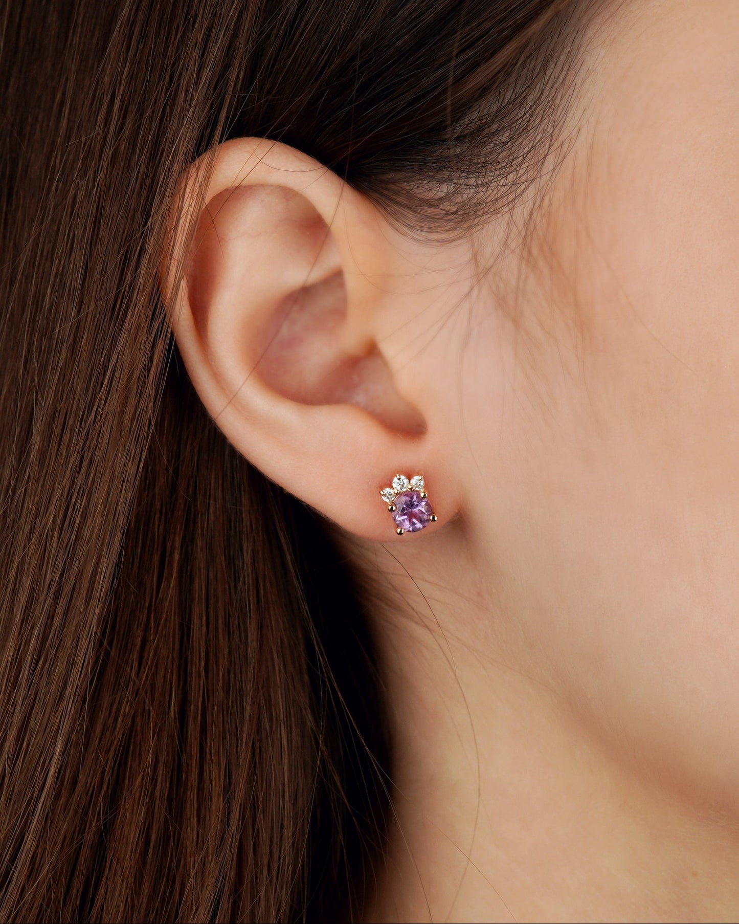 A close-up of a person's ear wearing a small stud earring with a purple heart-shaped gemstone and a silver-toned floral setting-delicate earrings-feminine jewelry- gold jewelry gift- amethyst jewelry