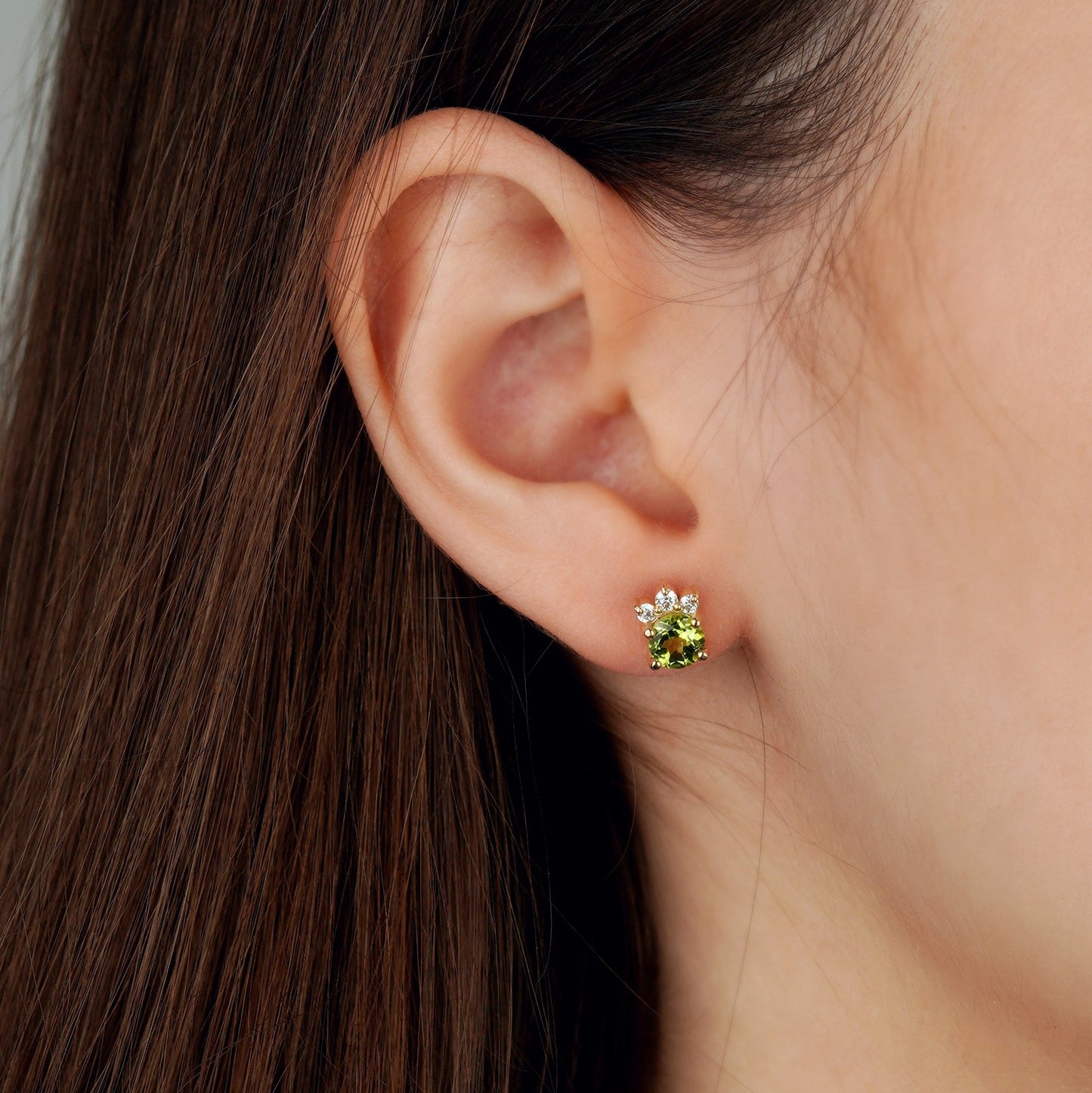 A close-up of a person's ear wearing a small flower-shaped stud earring with a green center and white petals in a gold-tone setting-, green gemstone earrings-sparkling earrings