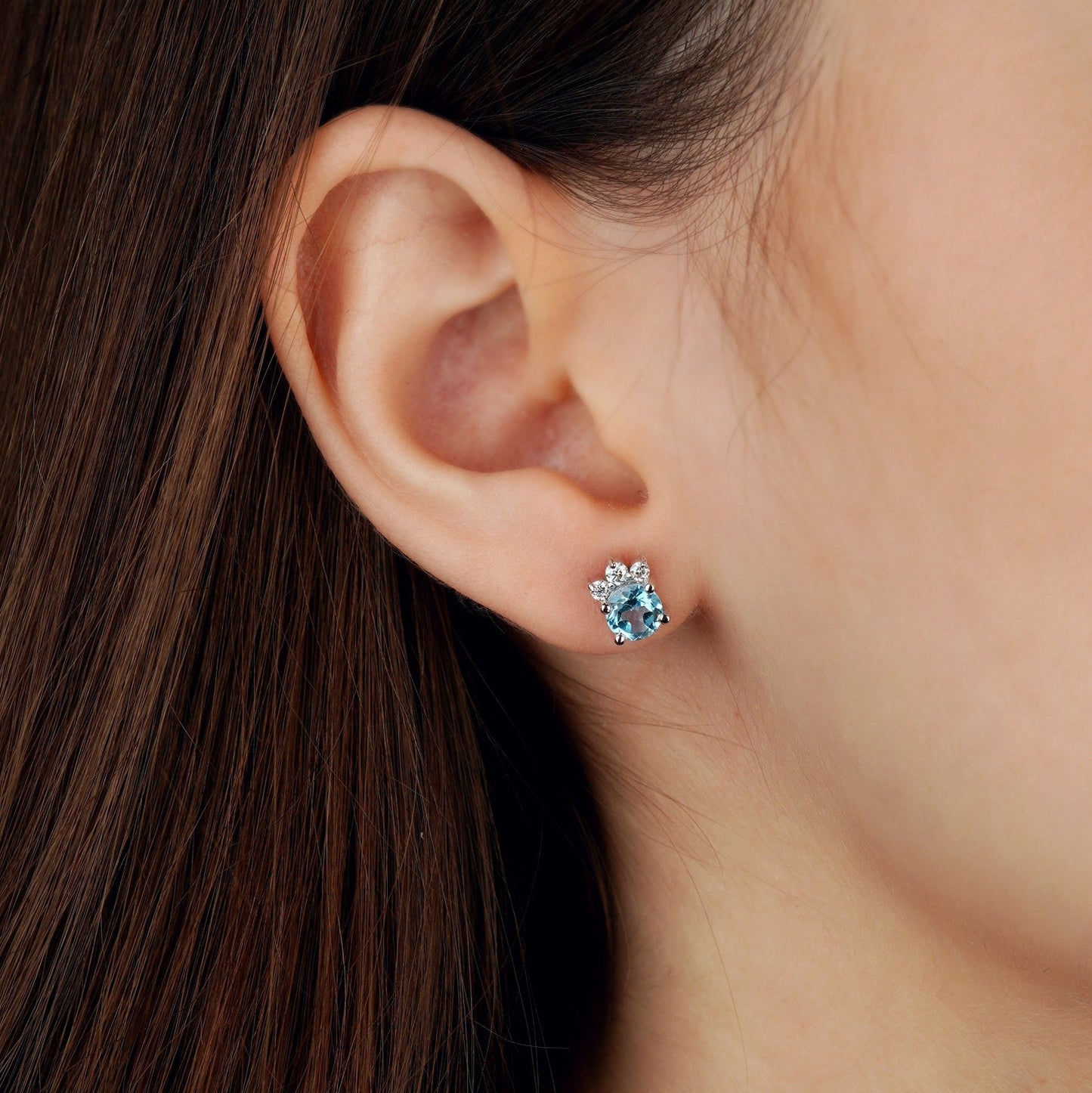 A close-up of a person's ear wearing a small floral stud earring with a light blue center stone and clear surrounding stones-feminine jewelry-gold jewelry gift-topaz jewelry- moissanite jewelry