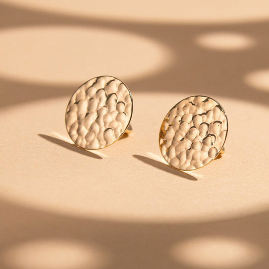 A pair of round gold-toned stud earrings with a textured, hammered surface, displayed on a beige surface with soft shadows-minimalist gold earrings-textured earrings