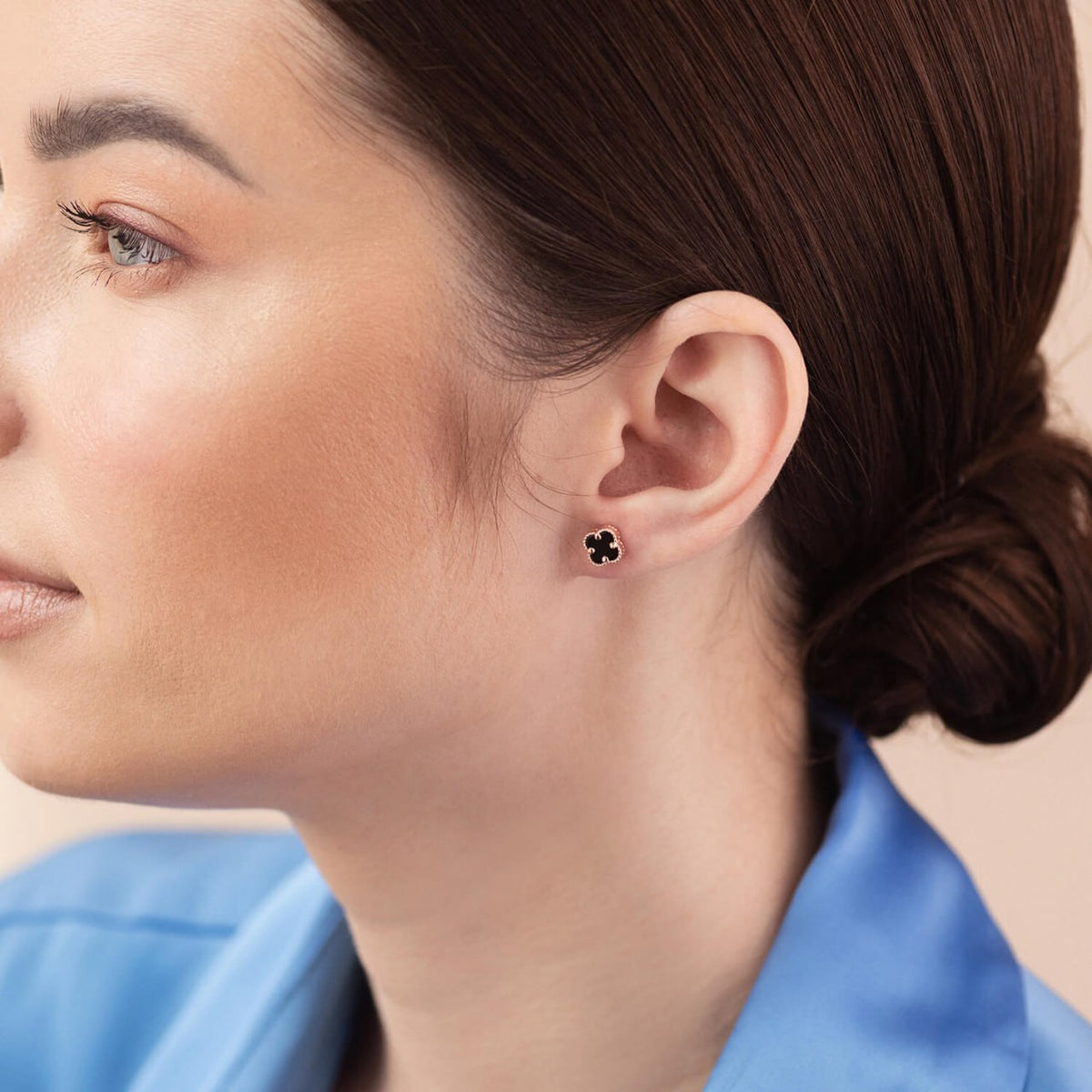 A close-up side profile of a woman with dark brown hair in a bun, wearing a small black clover stud earring with a silver-tone setting and a blue satin shirt-Metal: 14k Rose Gold (585)
Stone: Black Onyx
Diameter: 0.7 cm
Weight: ≈1.9 g