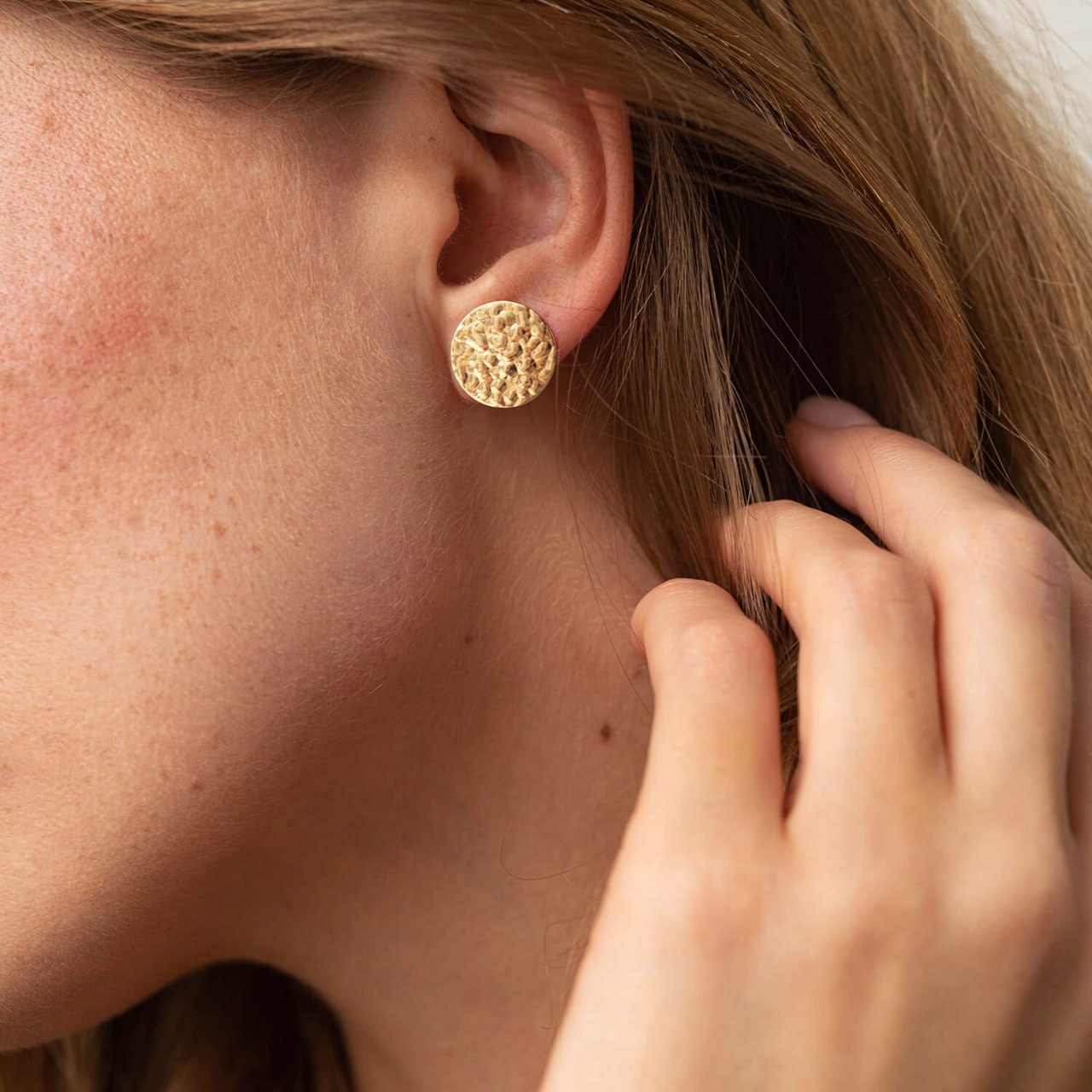 A close-up of a woman's ear wearing a textured gold stud earring, with her hand near her hair against -circle earrings-everyday gold earrings-gift for her