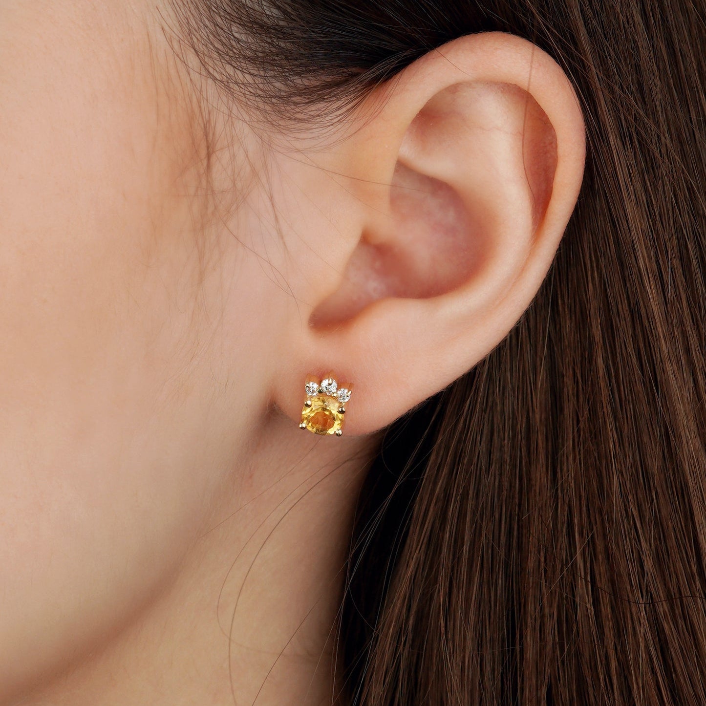 A close-up of a model's ear wearing a small gold stud earring with a yellow flower-like center and clear stones around it-Sunny Gemstone Stud Earrings