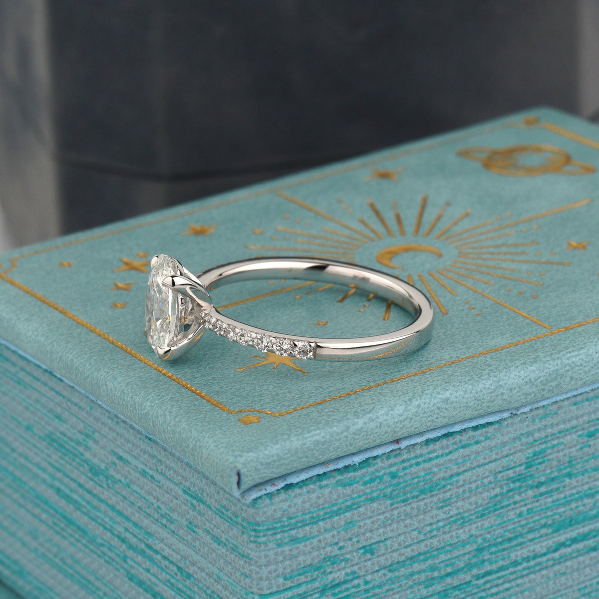 A white gold ring with a large clear rectangular gemstone and small side stones, displayed on a textured turquoise jewelry box with gold celestial designs-Setting: Classic prong with pavé band