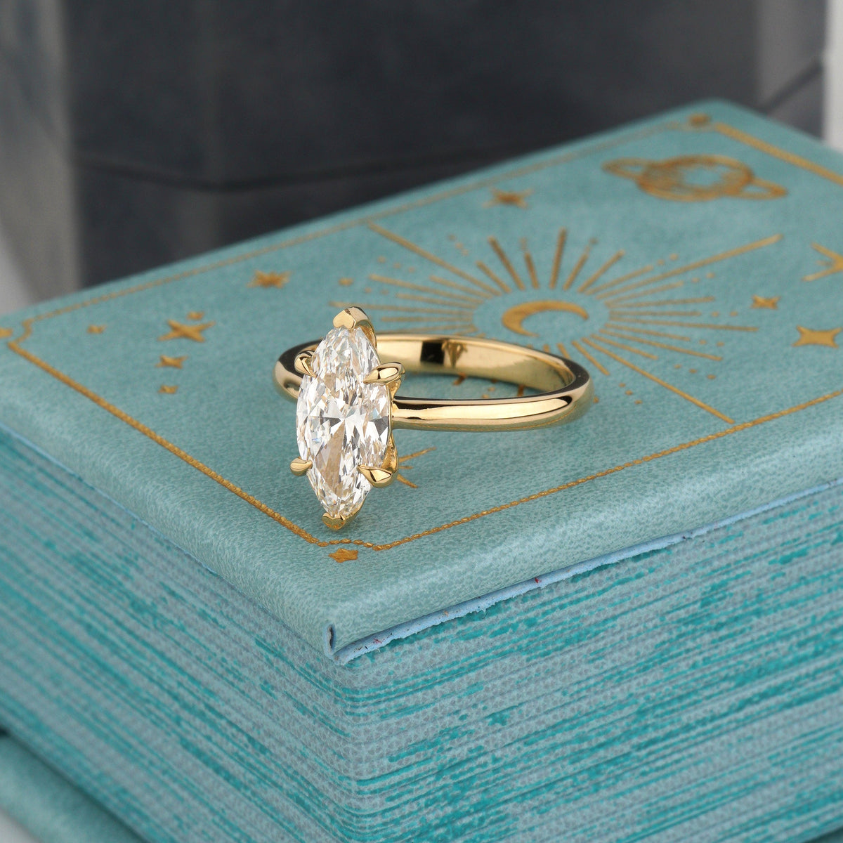 A gold ring with a marquise-cut clear gemstone in a prong setting, displayed on top of a textured turquoise gift box with gold celestial designs-proposal ring
lab diamond engagement ring
classic solitaire ring