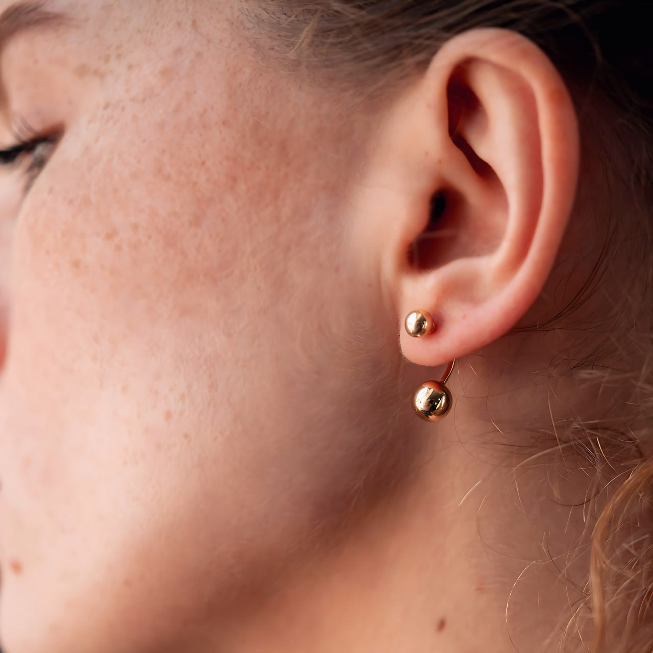A close-up of a person's ear wearing a pair of small gold stud earrings with a drop design- smooth, mirror-polished surface 