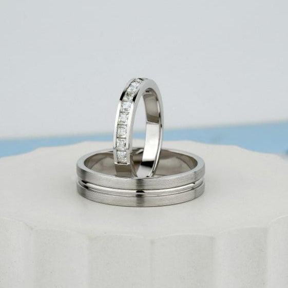 Two silver rings displayed on a white ceramic pedestal against a light gray background. One ring is plain, and the other has a row of small clear stones along the band.
