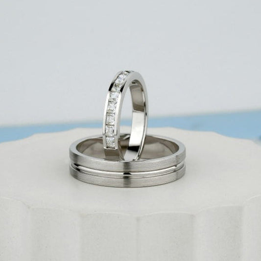 Two silver rings displayed on a white ceramic pedestal against a light gray background. One ring is plain, and the other has a row of small clear stones along the band.