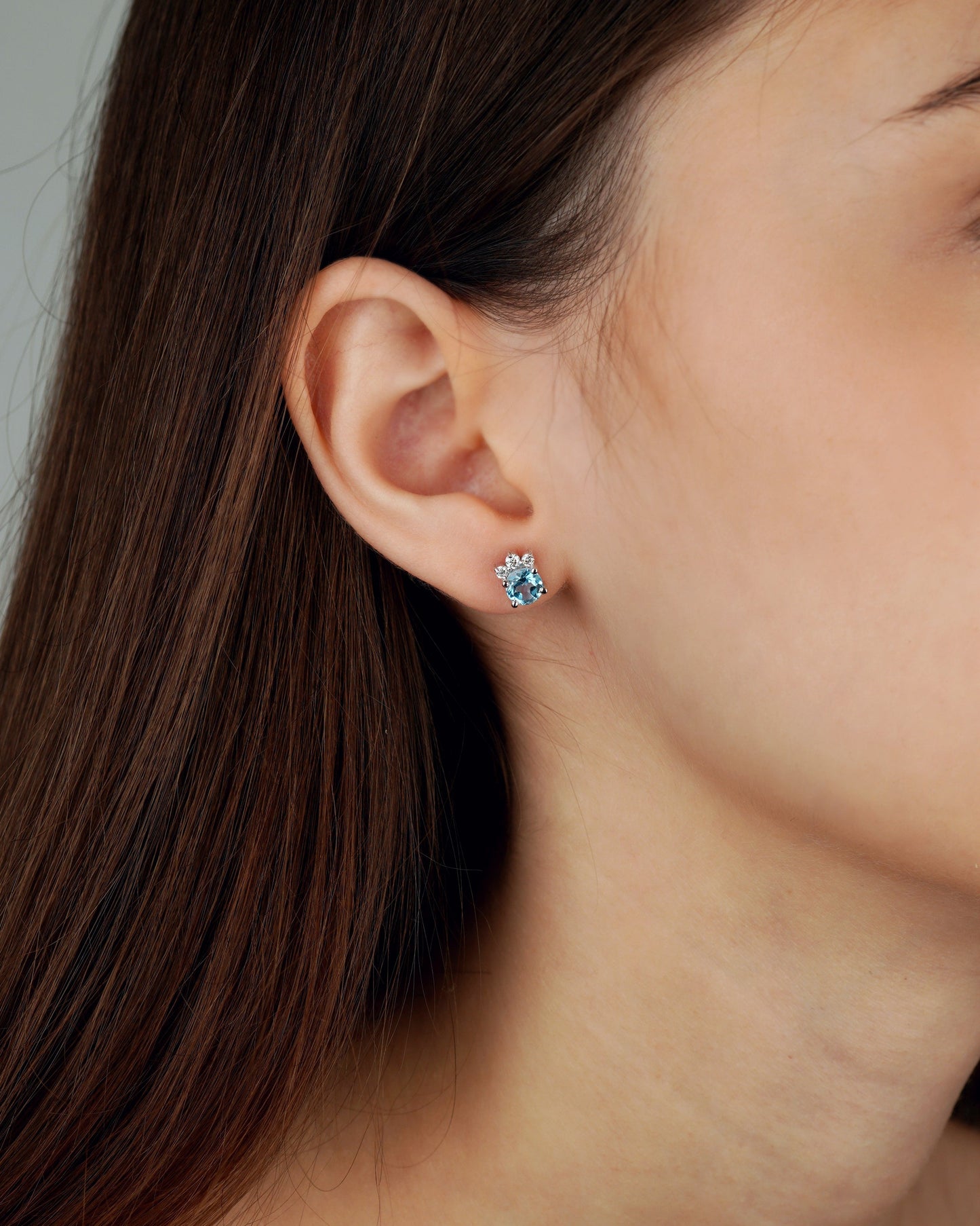A close-up of a woman's ear wearing a small stud earring with a light blue flower-like center and silver-toned petals-topaz earrings, moissanite earrings, swiss blue topaz earrings