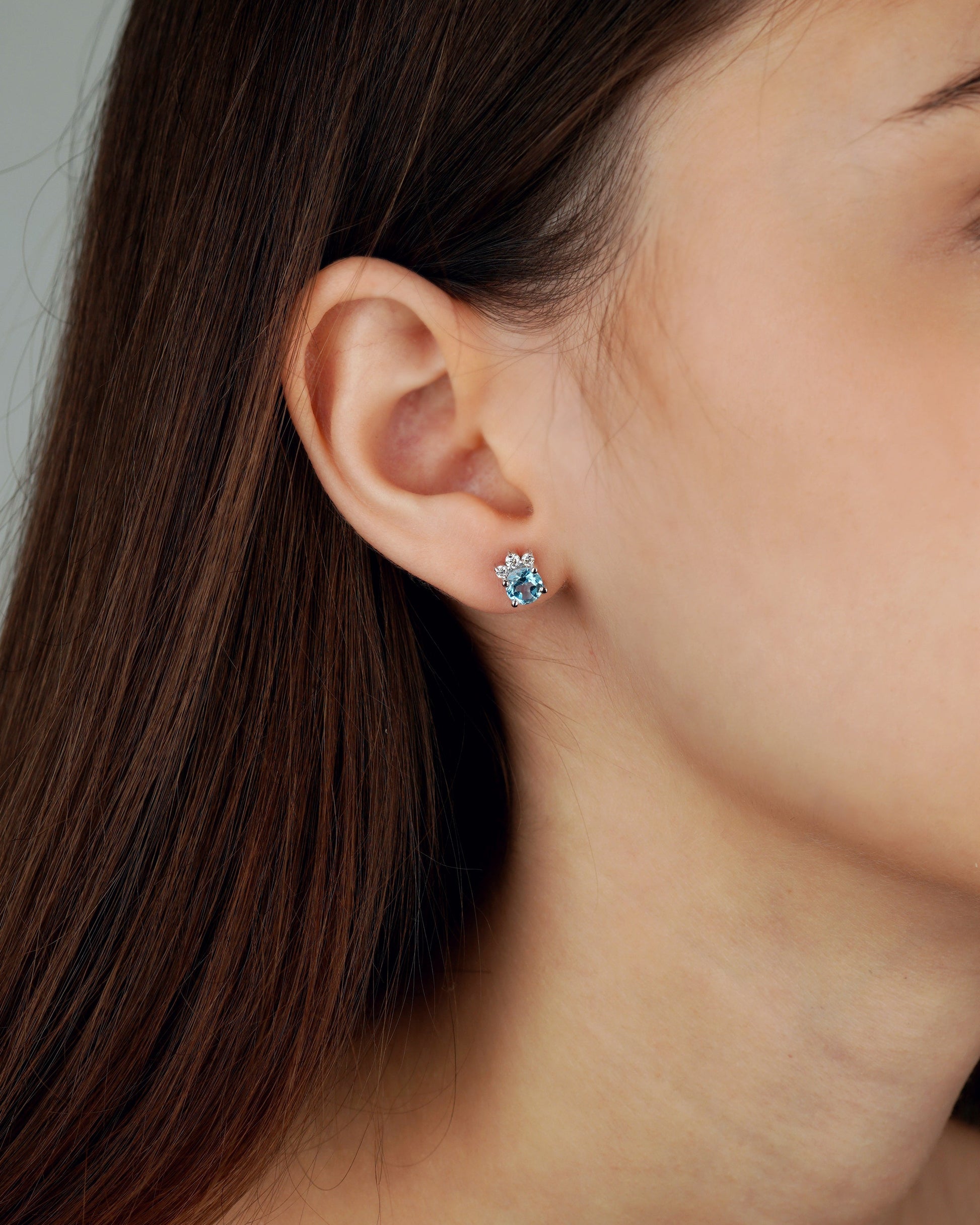 A close-up of a woman's ear wearing a small stud earring with a light blue flower-like center and silver-toned petals-topaz earrings, moissanite earrings, swiss blue topaz earrings