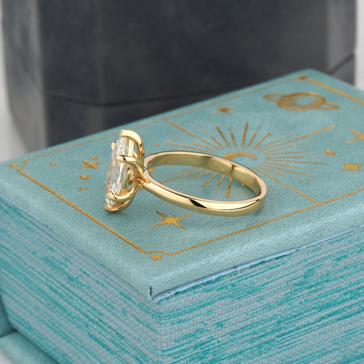 A gold ring with a clear gemstone displayed on top of a textured turquoise book-style box with gold celestial illustrations, with a dark box blurred in the background-lab diamond-engagement ring-classic solitaire ring-elongated diamond ring