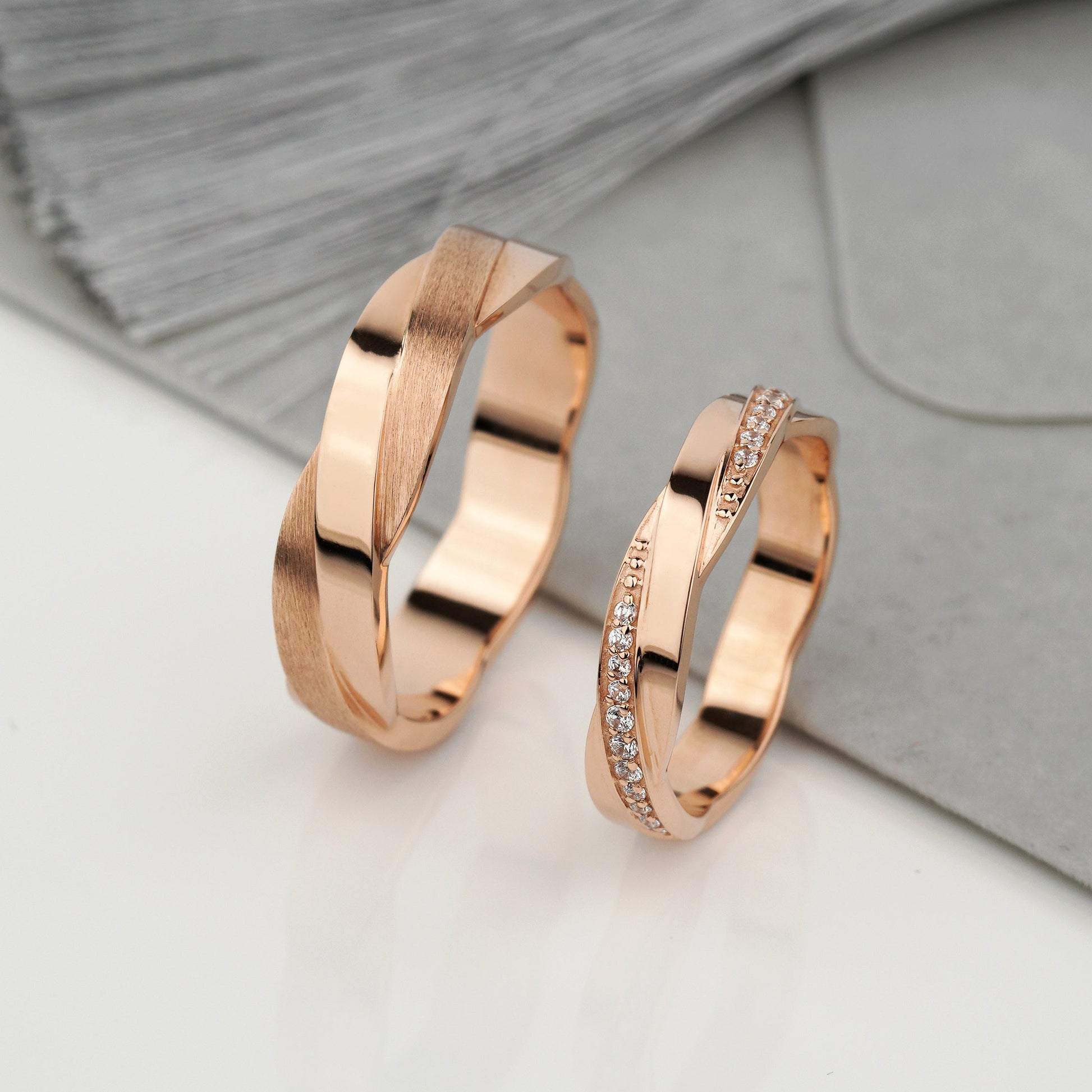 Two rose gold rings displayed on a white surface with a gray textured fabric background. One ring features a twisted, polished band design, and the other has a similar twisted band embellished with small clear stones.