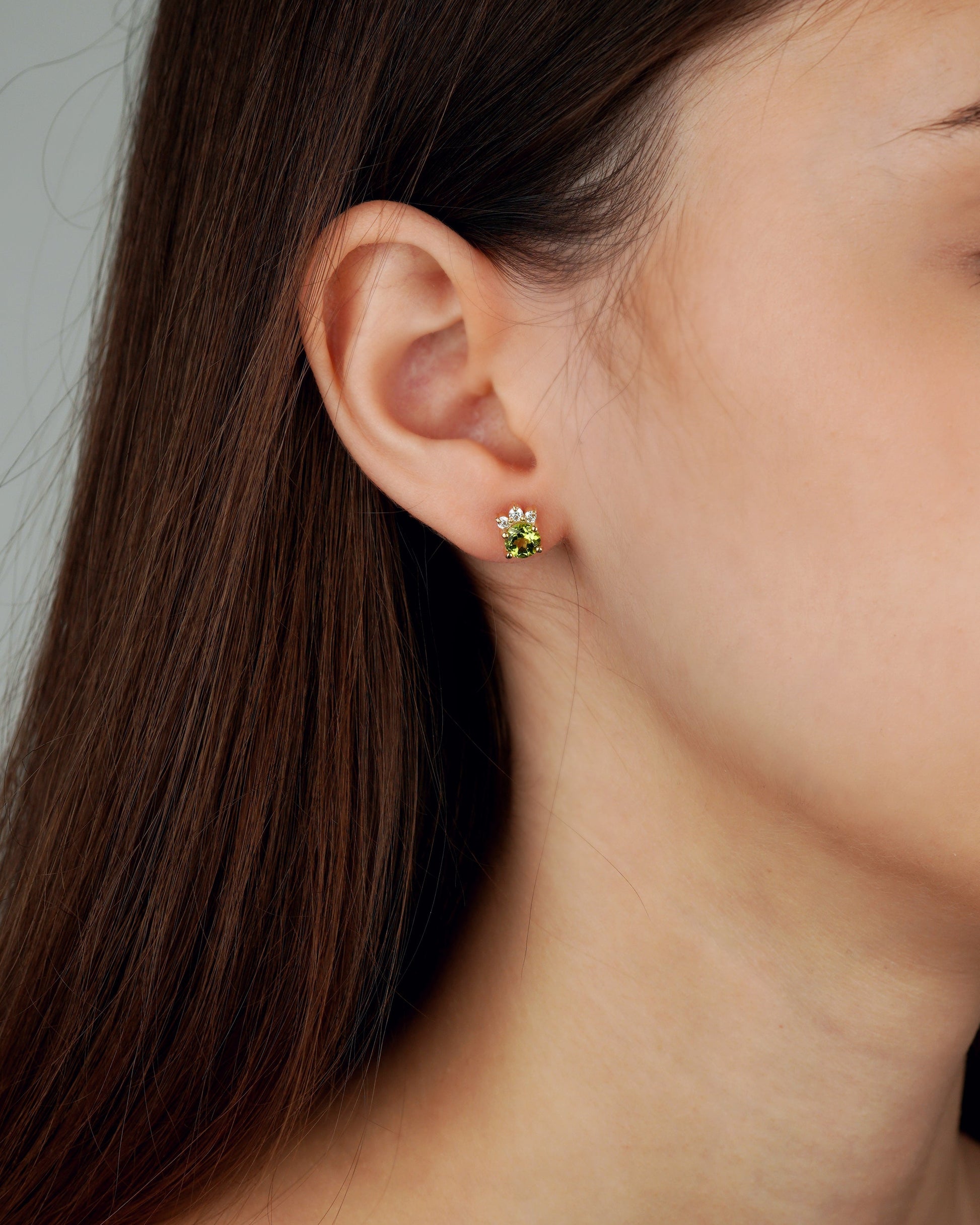 A close-up of a woman's ear wearing a small flower-shaped stud earring with a white center and green petals on a gold-tone setting-feminine jewelry- gold jewelry gift-peridot jewelry
