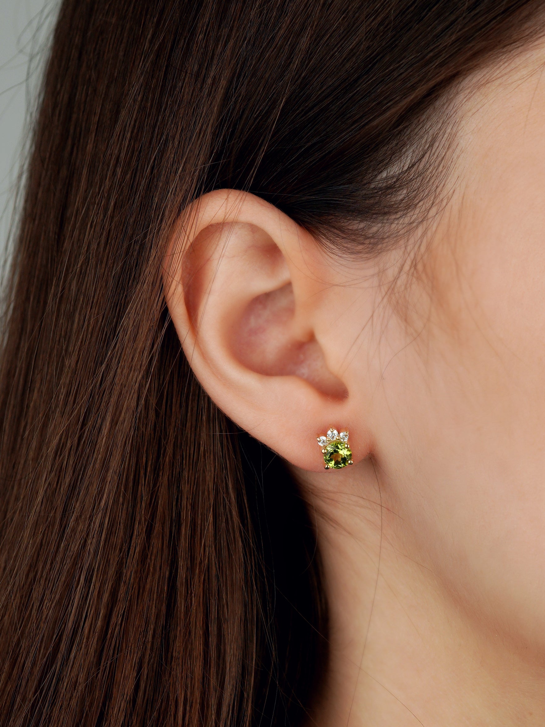 A close-up of a person's ear wearing a small flower-shaped stud earring with a green center and white petals in a gold-tone setting-, green gemstone earrings-sparkling earrings