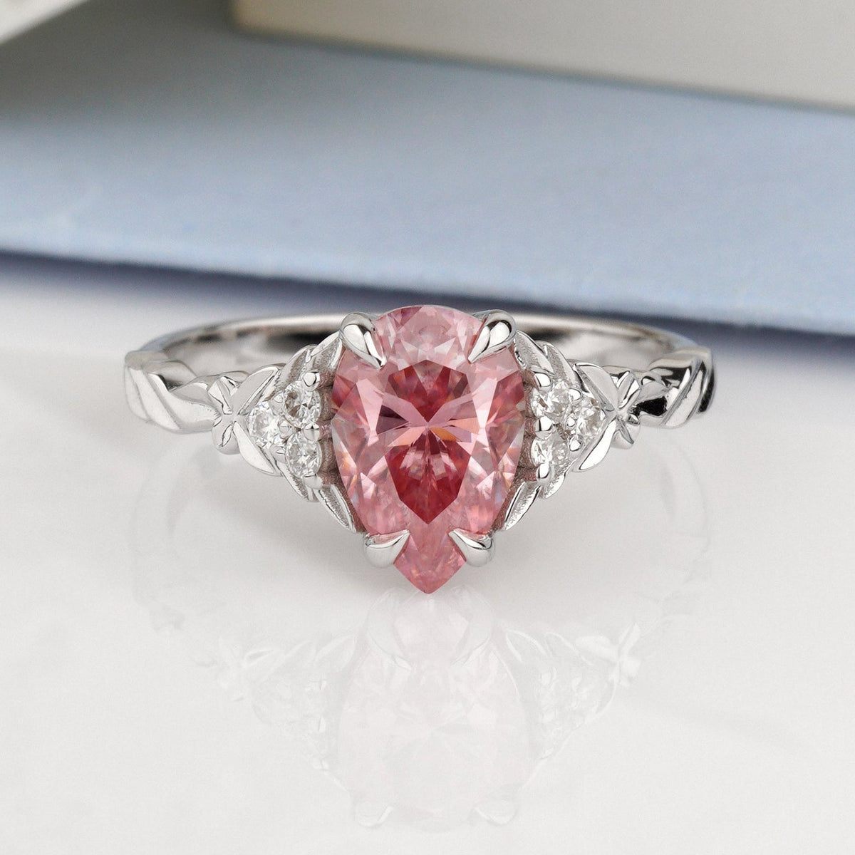 A silver ring featuring a heart-shaped pink gemstone in a prong setting, accented with small clear stones on either side of the band-pink moissanite ring-pear engagement ring-white gold engagement ring