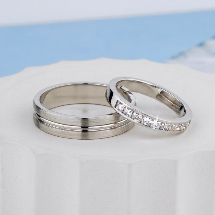 Two silver-toned wedding bands displayed on a white pedestal. One band is smooth and polished, while the other is adorned with a row of clear pavé-set stones-Women's band: 3 mm wide