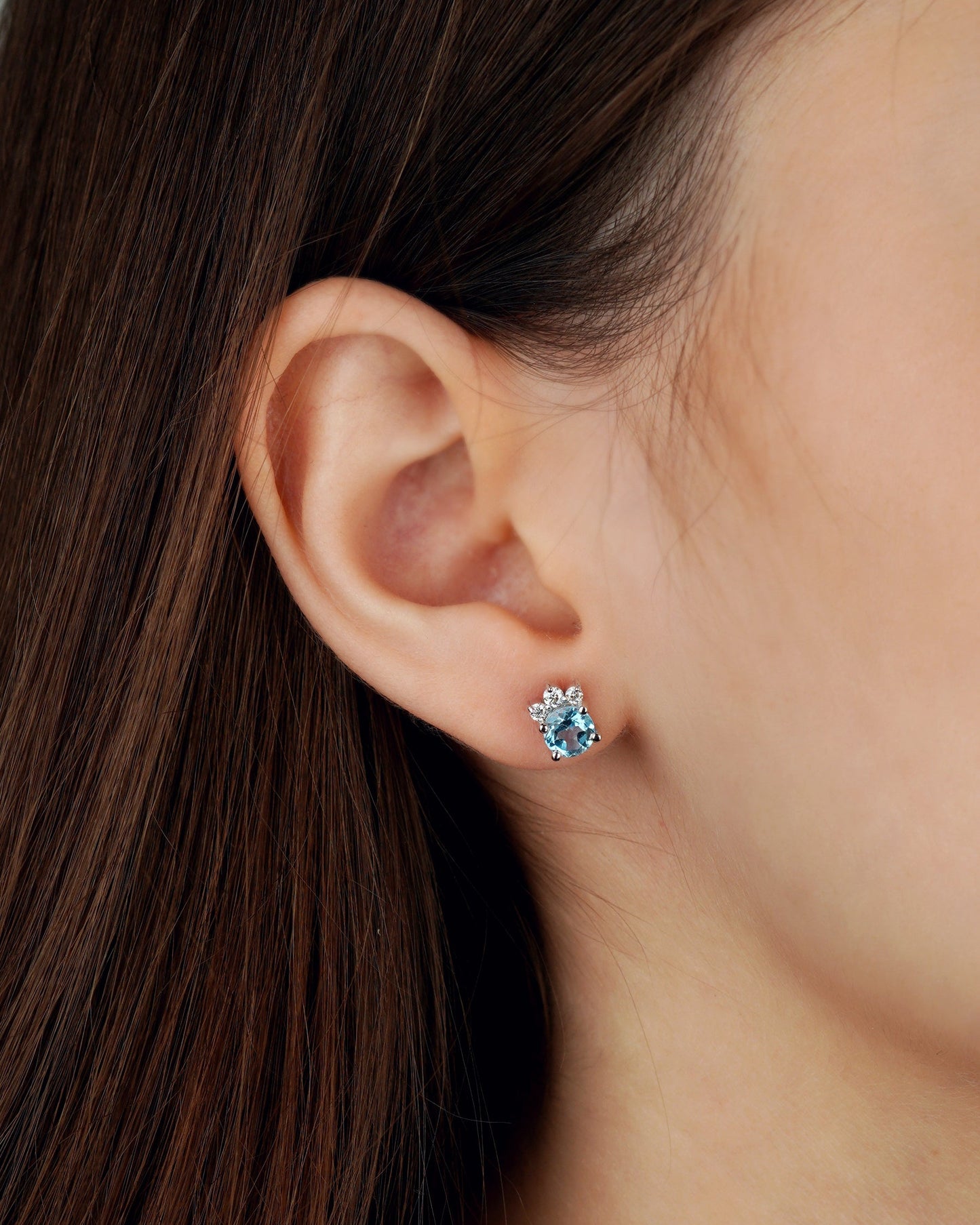 A close-up of a person's ear wearing a small floral stud earring with a light blue center stone and clear surrounding stones-feminine jewelry-gold jewelry gift-topaz jewelry- moissanite jewelry