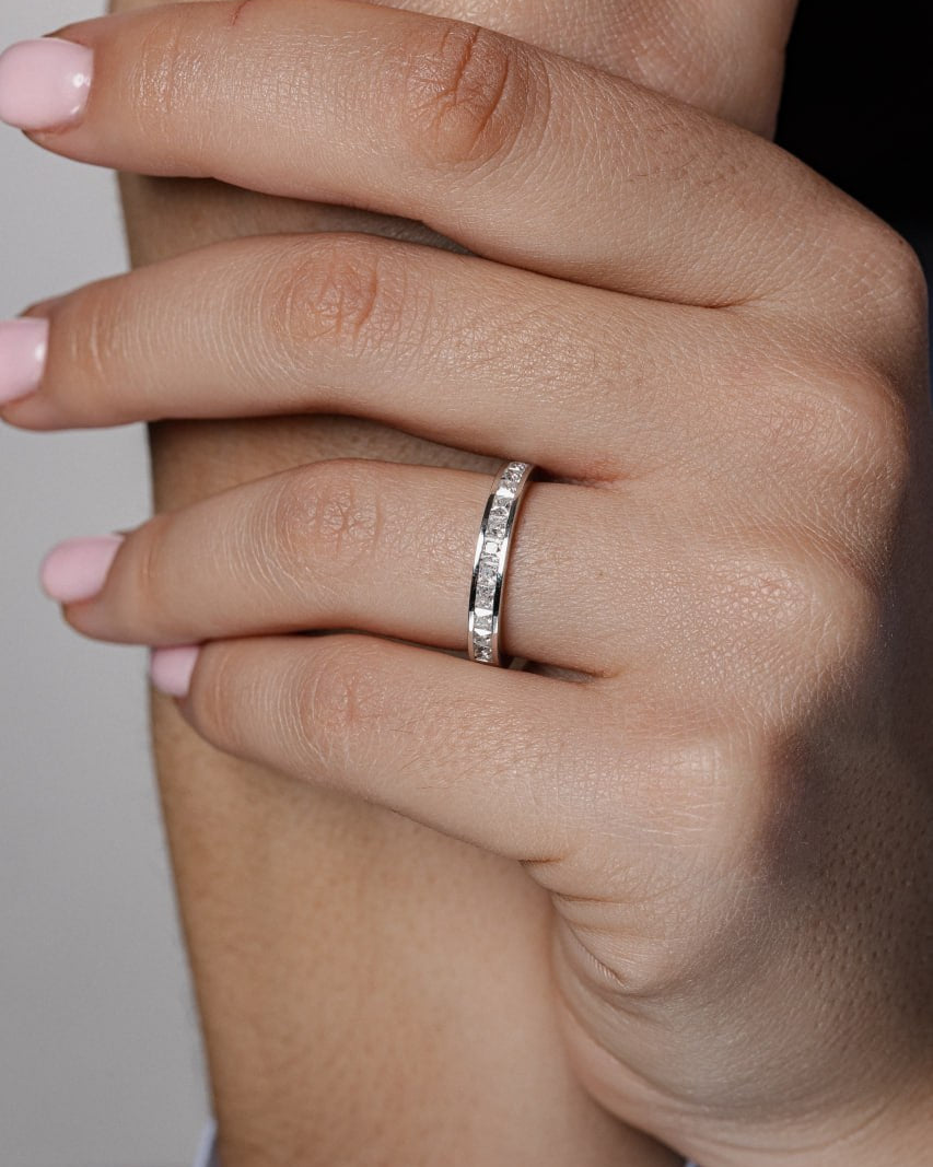 A close-up of a person's hands with light pink manicured nails, wearing a thin silver band ring with small clear stones on one finger- men's band with a polished groove pairs with a sparkling princess-cut channel-set women's band 