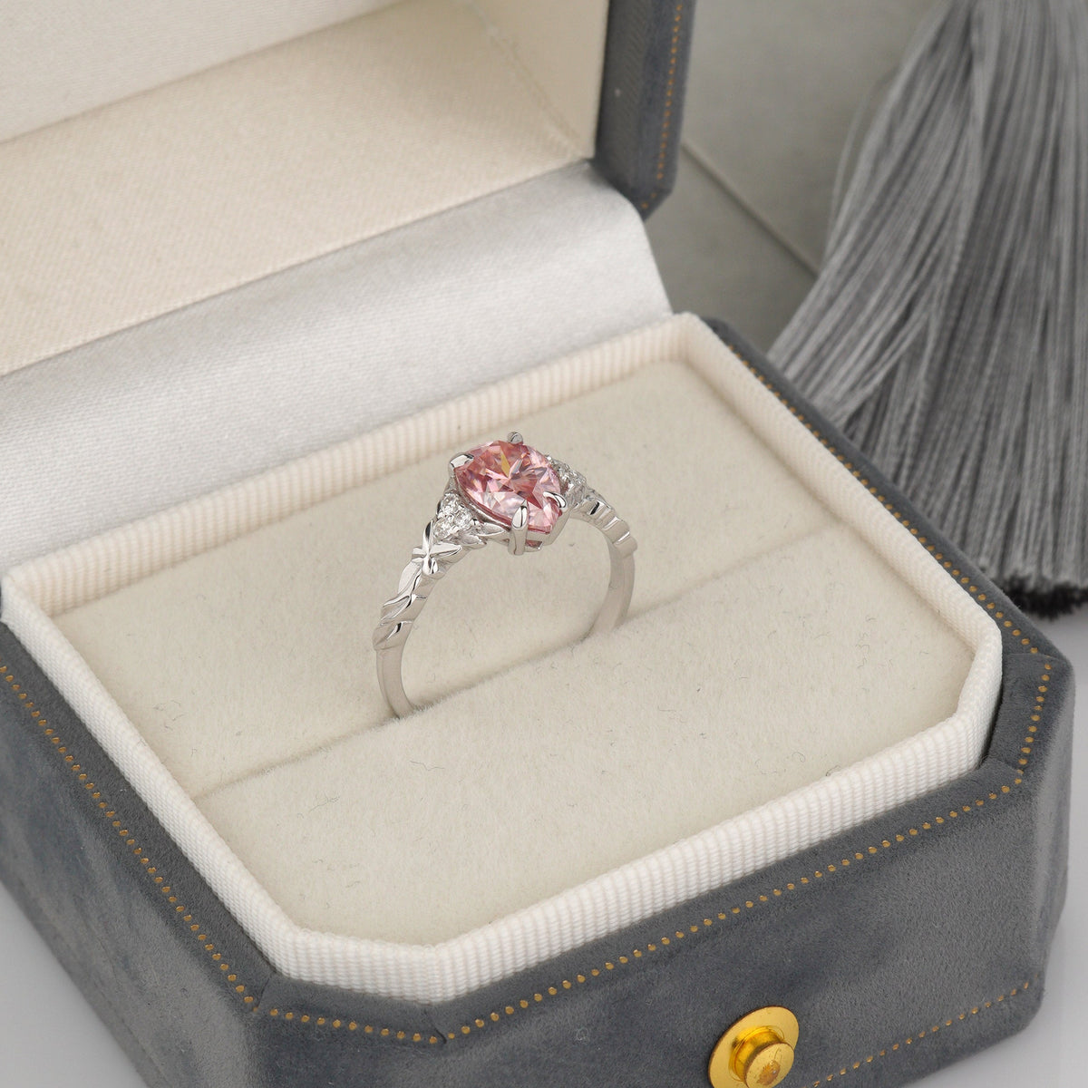 A silver ring with a heart-shaped pink gemstone is displayed inside an open gray jewelry box with a cream velvet insert-pink moissanite ring-pear engagement ring-white gold engagement ring-vintage promise ring