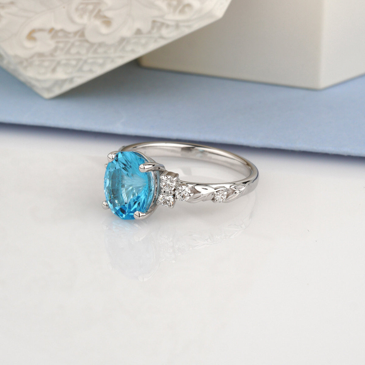 A white gold ring featuring an oval-cut light blue gemstone and small clear accent stones on a white surface, with a decorative white textured object and pale blue background in the upper portion-oval gemstone ring-topaz engagement ring-floral engagement ring 