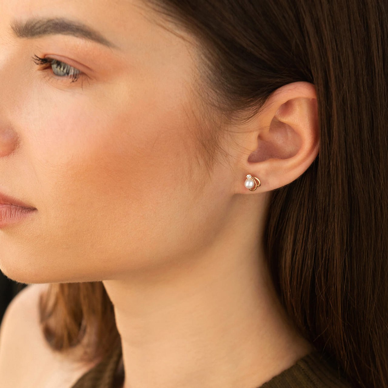 A close-up side profile of a woman wearing a small gold stud earring with a white pearl-like drop, shown against her ear and hair-rose gold earrings-14k gold pearl studs-delicate pearl earrings