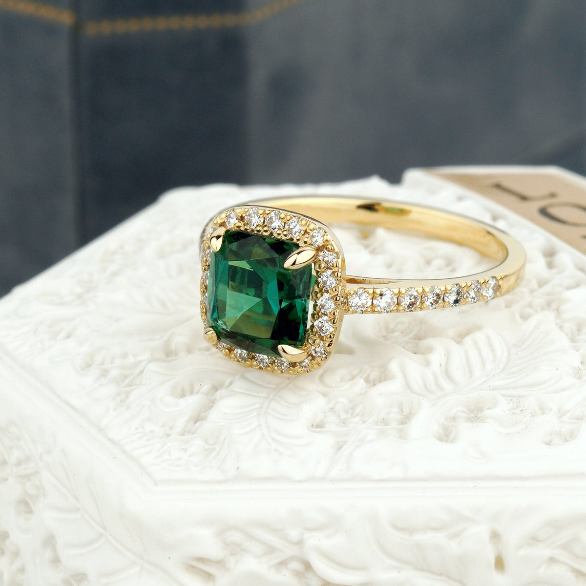 A yellow gold ring with a cushion-cut green gemstone and a halo of small clear stones, displayed on a white carved pedestal with a dark jewelry box in the background-yellow gold engagement ring-unique engagement ring-green gemstone ring-alternative engagement ring