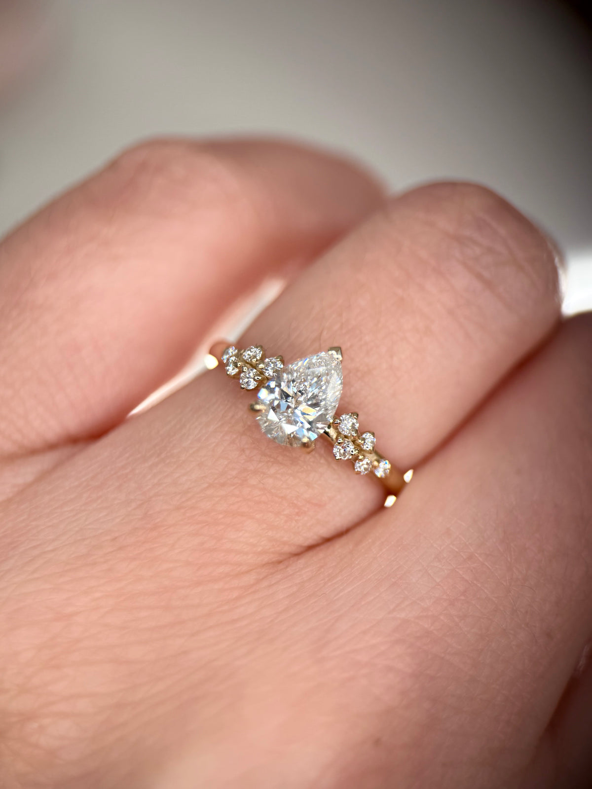A close-up of a hand wearing a yellow gold ring with a pear-cut center stone and small accent stones along the band-Side Stones: Round Lab-Grown Diamonds, 0.11 ct total-Setting: Cluster side accents
