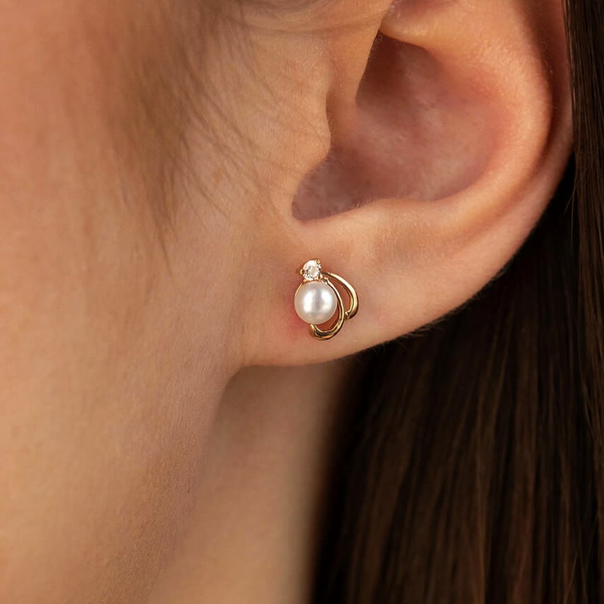 A close-up of a person's ear wearing a small gold hoop earring with a white pearl and a tiny clear stone at the top-minimalist gold earrings-solid gold jewelry-everyday pearl studs