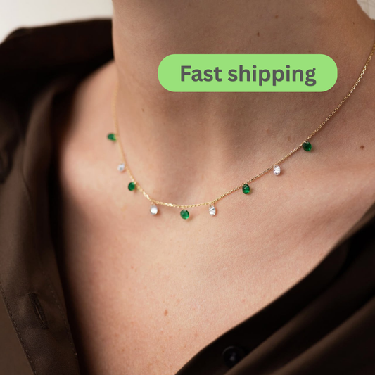 A close-up of a model's neck and chest wearing a delicate gold necklace with small green and clear gemstone accents