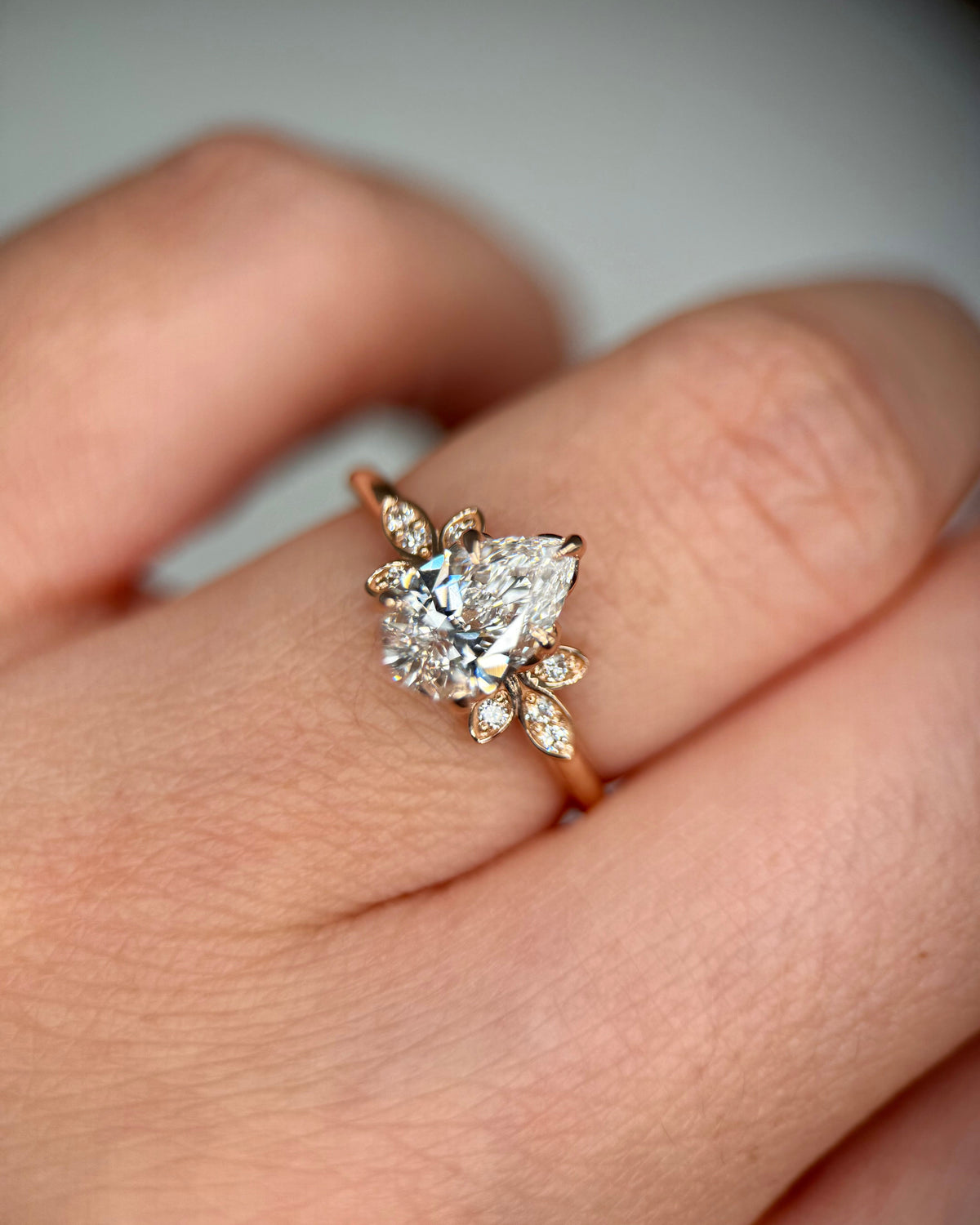 A close-up of a hand wearing a rose gold ring with a pear-cut diamond center stone and smaller marquise-cut diamonds in a floral cluster setting-vintage engagement ring -unique engagement ring-proposal ring-floral halo engagement ring