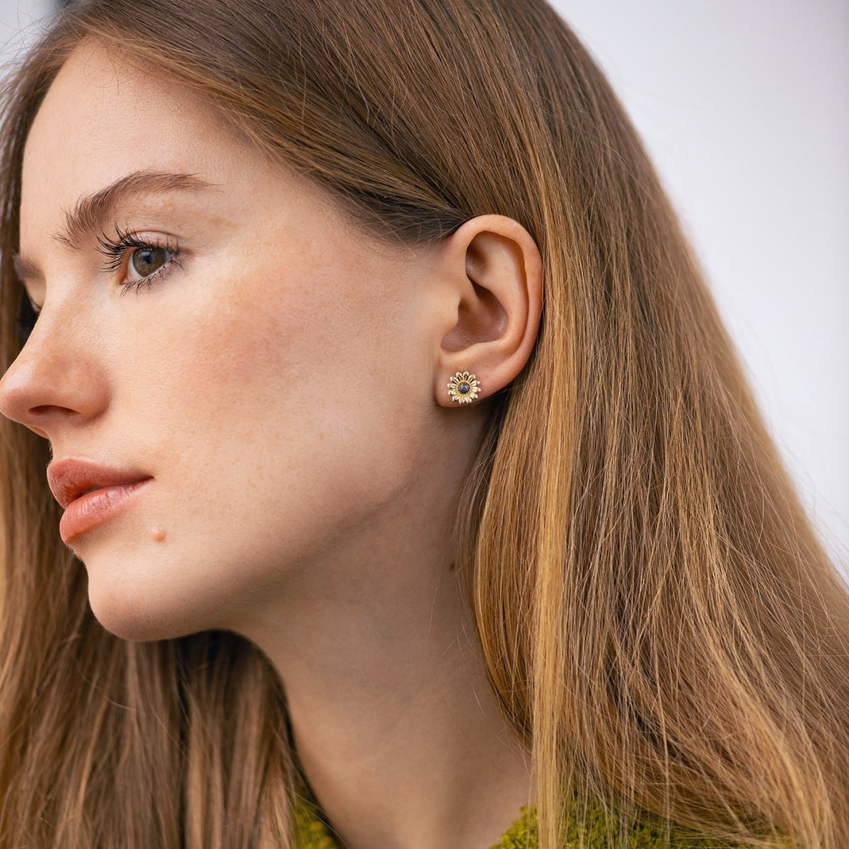 A close-up side profile of a woman with long light brown hair wearing a small gold flower-shaped stud earring with tiny clear stones-nature inspired earrings-sunflower gold studs-everyday earrings