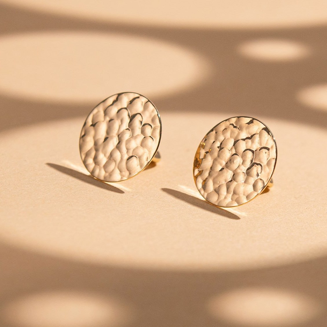 A pair of round gold-toned stud earrings with a textured, hammered surface, displayed on a beige surface with soft shadows-minimalist gold earrings-textured earrings