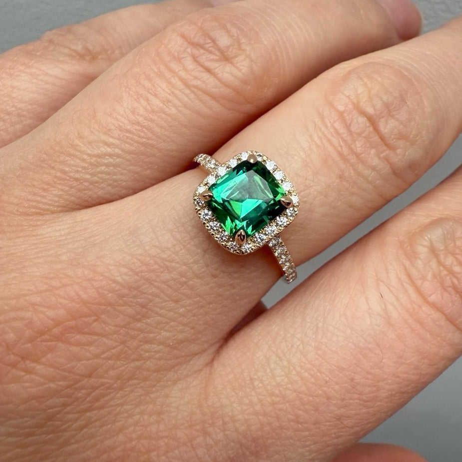 A close-up of a hand wearing a ring with a square-cut green gemstone surrounded by a halo of small clear stones on a silver-toned band-Center Stone: Natural Green Tourmaline, Cushion Cut, 1.7 ct
Accent Stones: Natural Diamonds, VVS2/F, 0.41 ct total