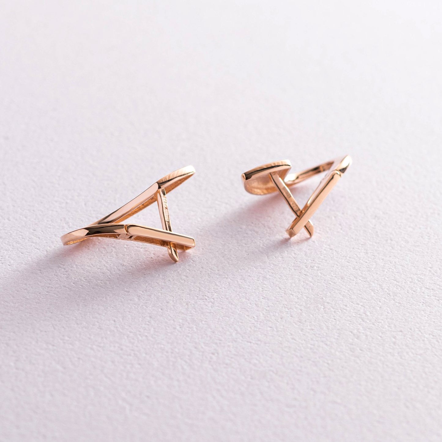 A pair of rose gold geometric stud earrings with a twisted, abstract triangular design, displayed on a clean, light pink textured surface-Earring height: 2.2 cm-Finish: High polish