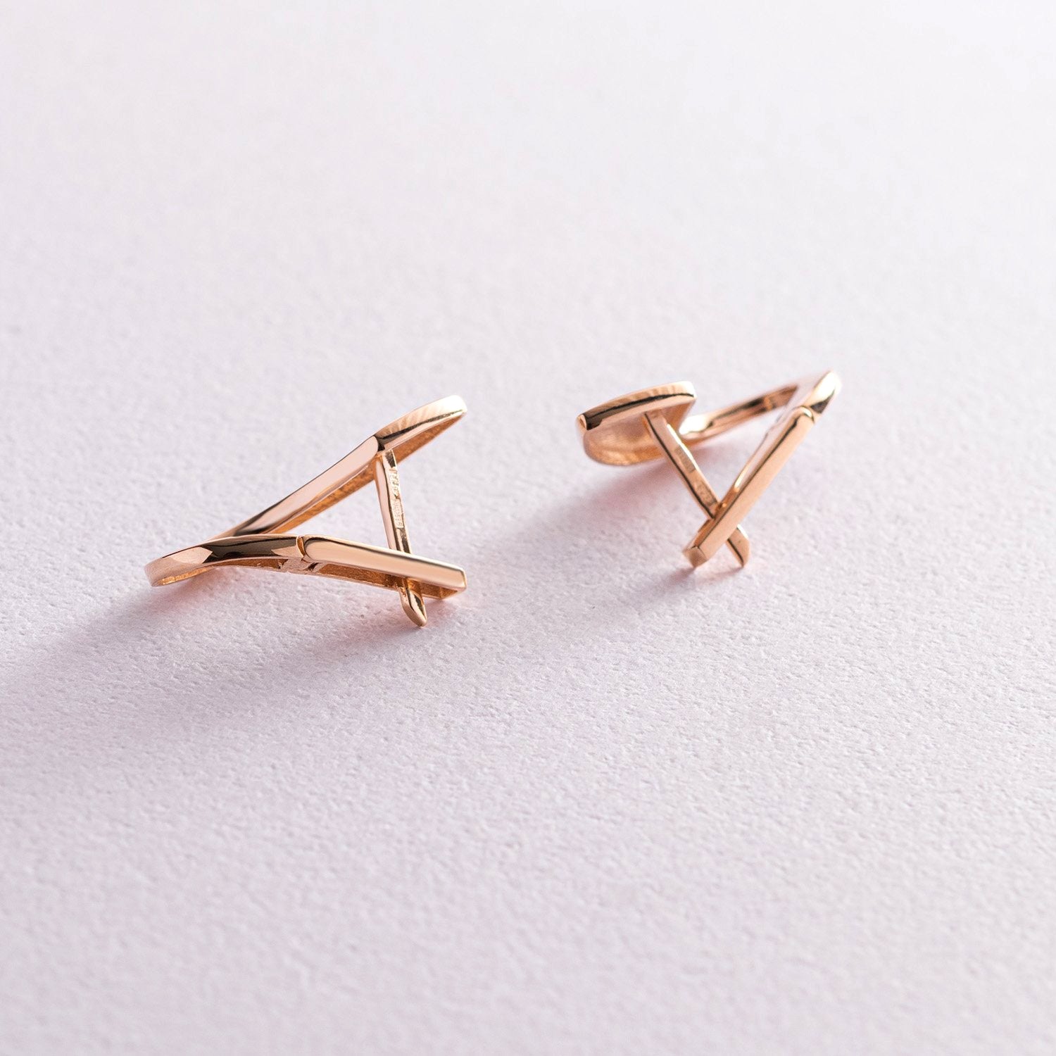 A pair of rose gold geometric stud earrings with a twisted, abstract triangular design, displayed on a clean, light pink textured surface-Earring height: 2.2 cm-Finish: High polish