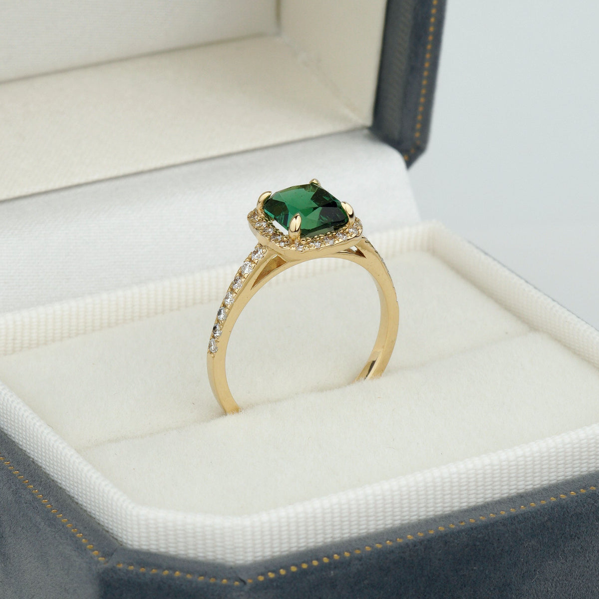 A gold ring with a square-cut green gemstone and small clear accent stones, displayed in an open dark gray jewelry box with a white cushion-unique engagement ring
green gemstone ring
alternative engagement ring
solid gold ring