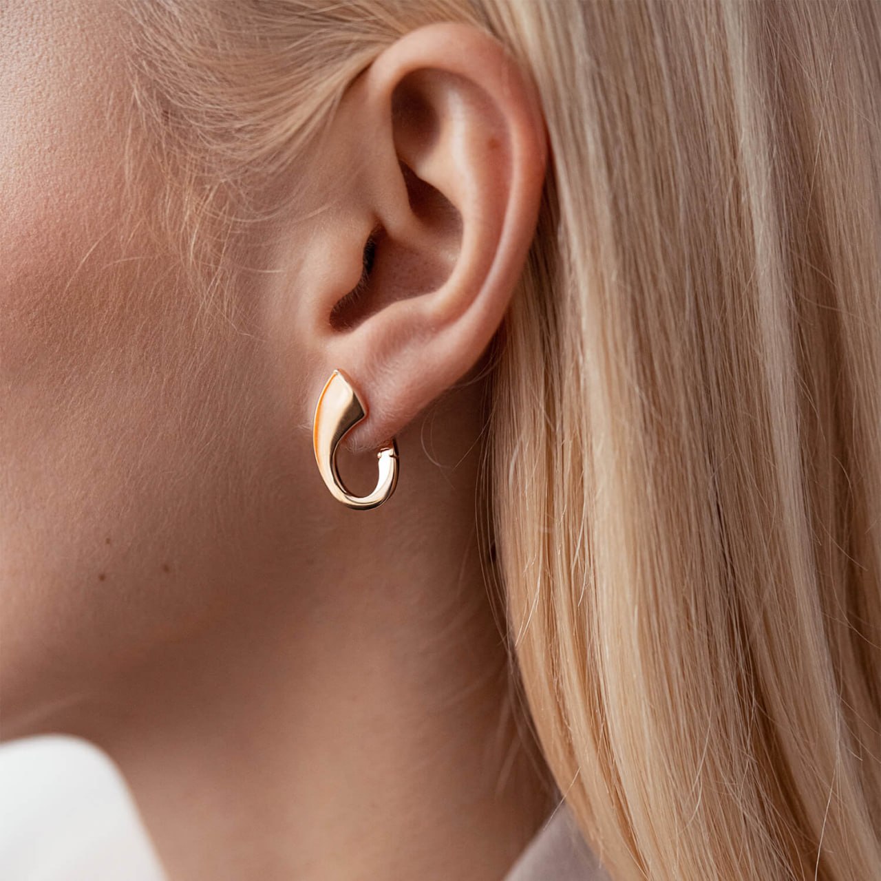A close-up of a model's ear wearing a small, polished gold huggie hoop earring.