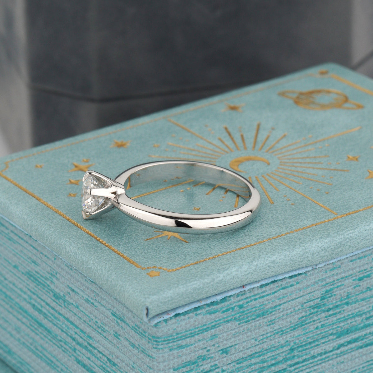A silver-toned solitaire ring with a clear triangular gemstone rests on a textured light blue jewelry box with gold celestial designs-lab grown diamond-promise ring for her-minimalist engagement ring