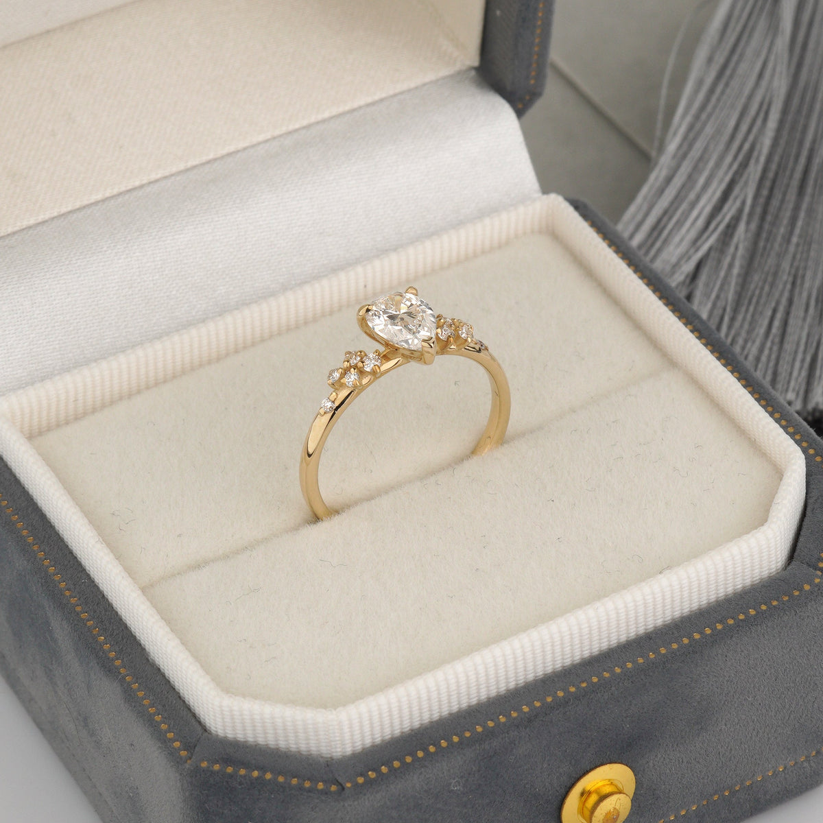 A gold ring with a heart-shaped clear gemstone and small side stones, displayed in an open gray jewelry box with a cream velvet interior.