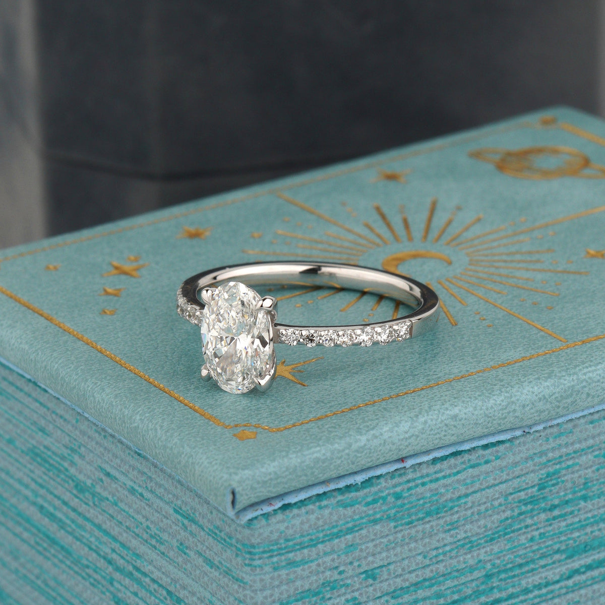 A white gold engagement ring with a round-cut diamond and a pavé band, resting on a textured turquoise gift box with gold celestial designs-Side Stones: Diamonds, 0.17 ct total