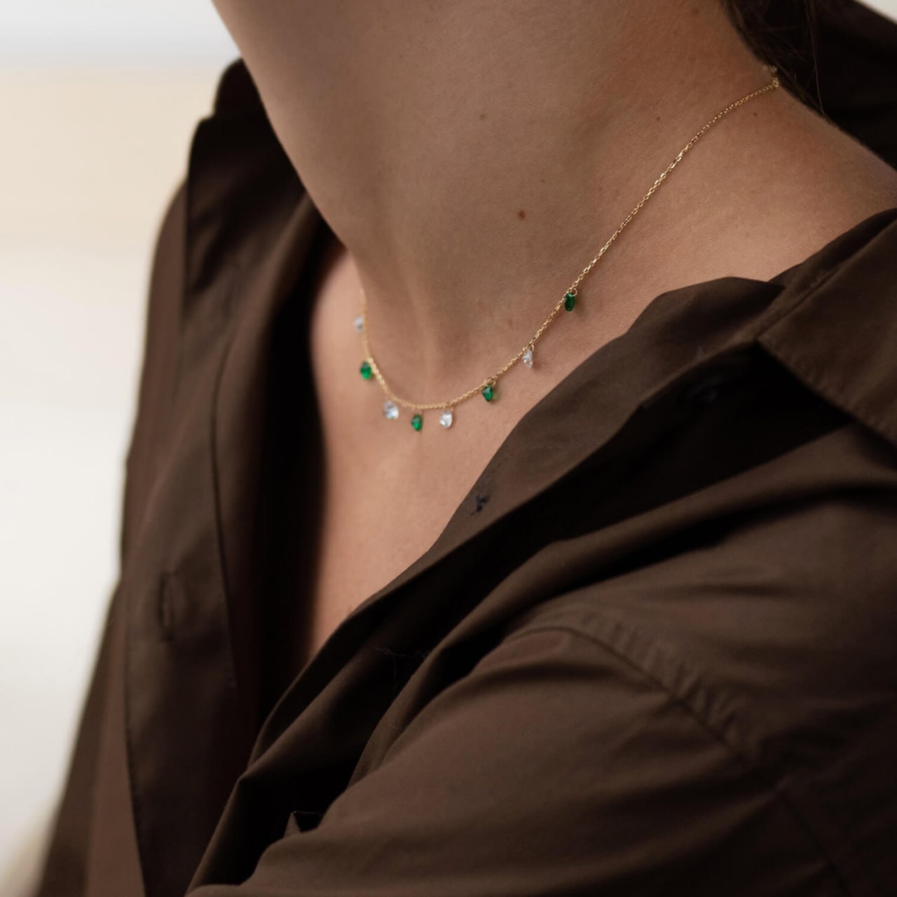 A close-up of a delicate gold chain necklace with small green and clear teardrop-shaped stones, worn by a person in a dark brown shirt-charm necklace-solid gold jewelry-Everyday gold necklace