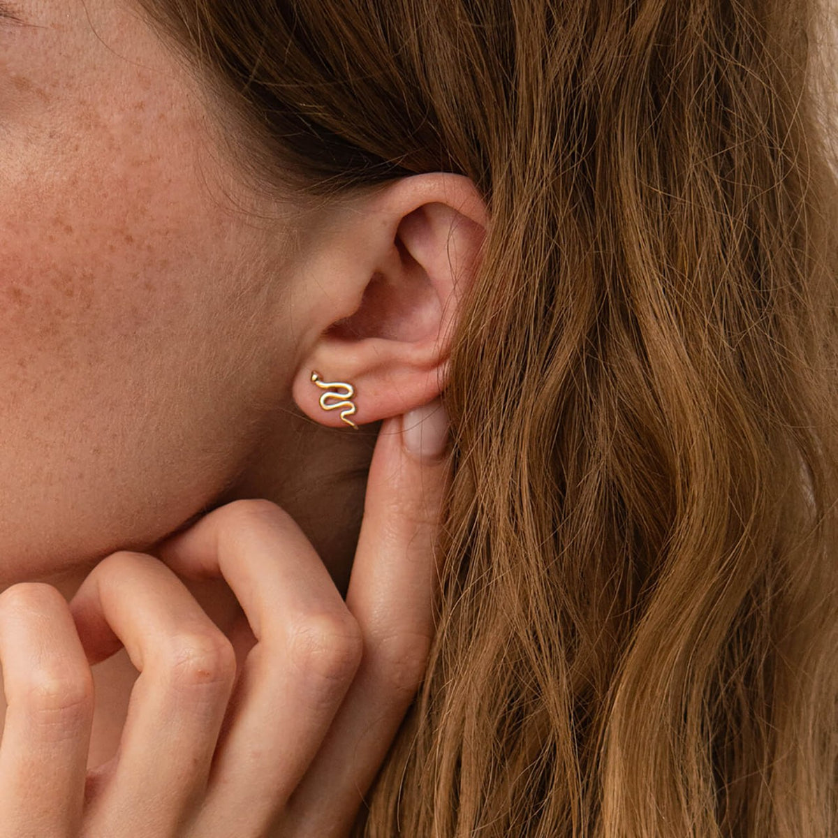 A close-up of a woman's ear wearing a delicate gold snake-shaped stud earring, with her hand resting near her chin.
