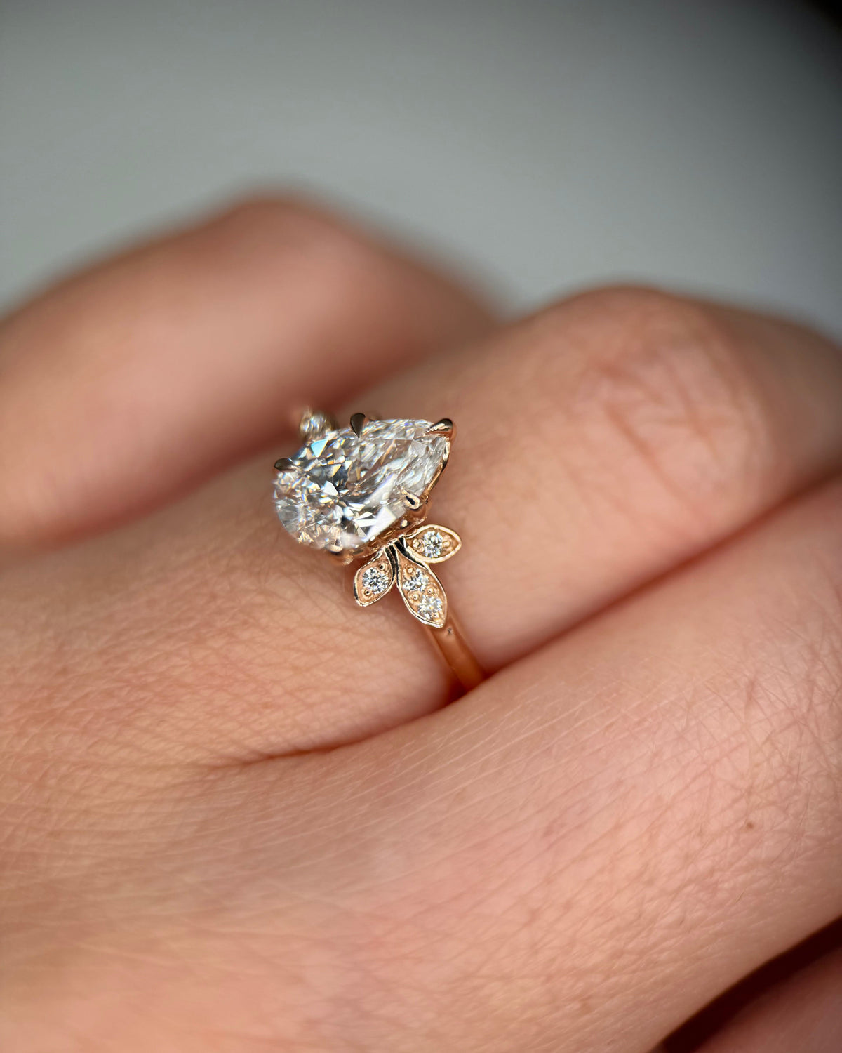 A close-up of a hand wearing a rose gold ring with a pear-cut clear center stone and small leaf-shaped side stones-unique engagement ring
proposal ring-floral halo engagement ring