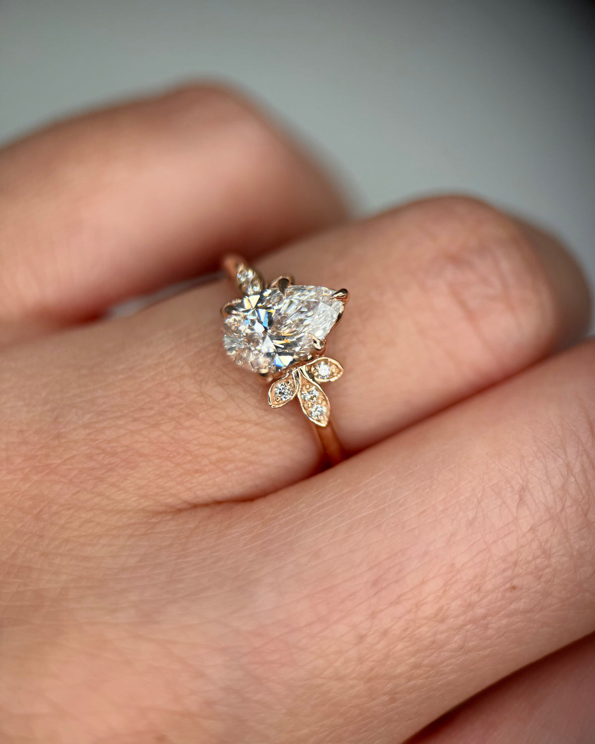 A close-up of a hand wearing a rose gold ring with a pear-cut diamond center stone and small diamond leaf accents-Side Stones: Diamonds, 0.08 ct total