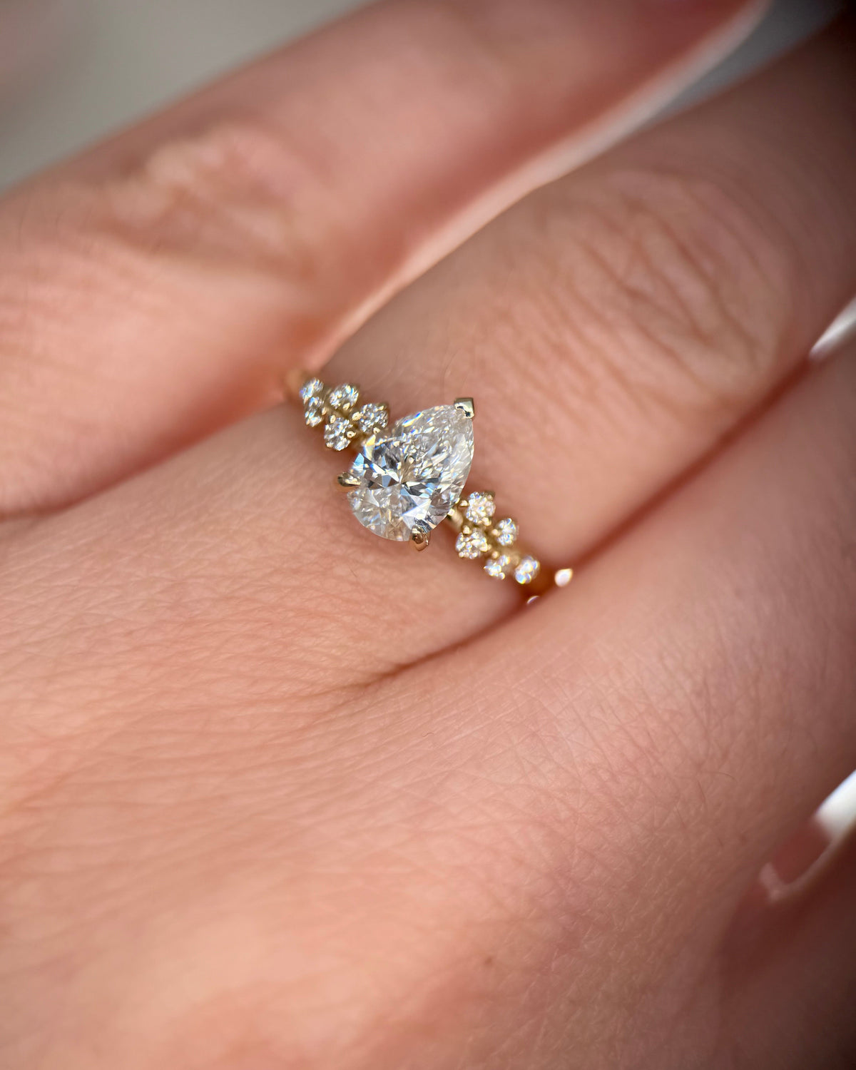 A close-up of a gold ring with a pear-cut center stone and small accent stones, worn on a finger-pear diamond ring-yellow gold engagement ring-lab diamond ring