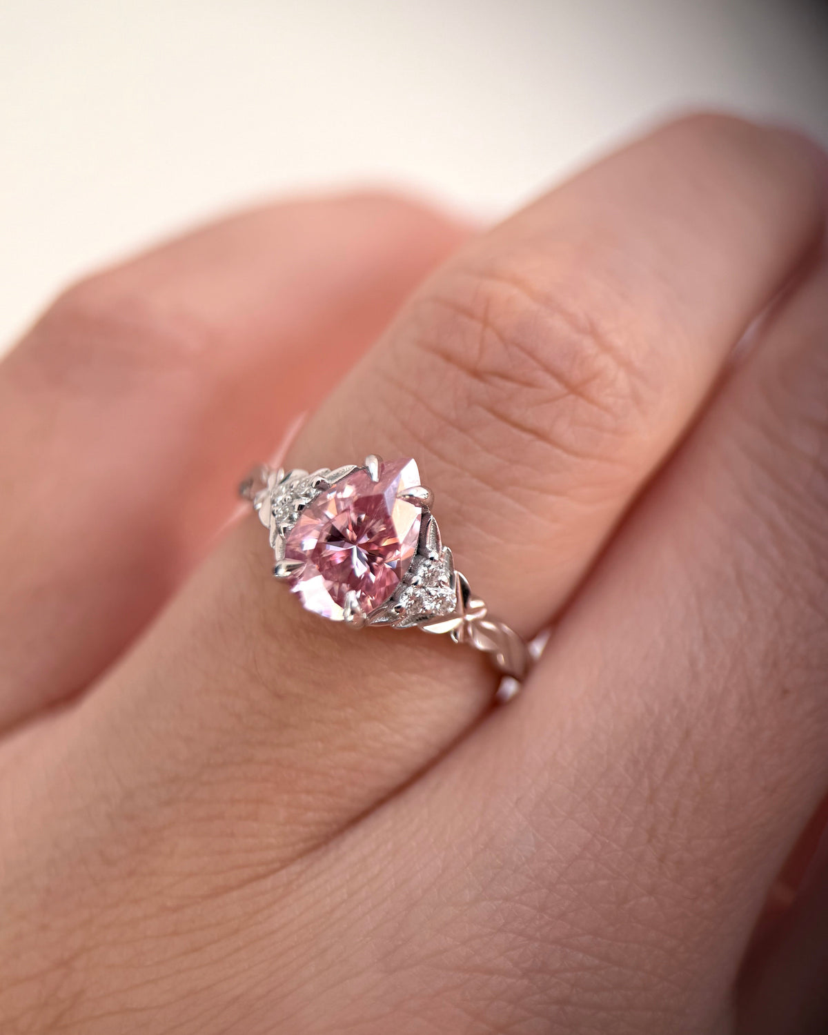 A close-up of a hand wearing a silver-toned ring with a prominent round pink gemstone and small accent stones on a soft light background-moissanite engagement ring-colored stone ring-unique engagement ring