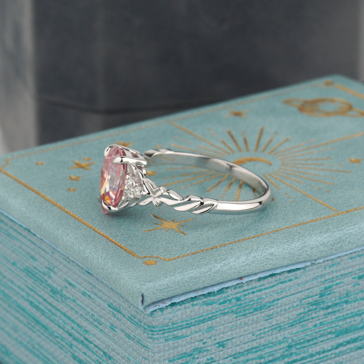 A white gold ring with a heart-shaped pink gemstone and a sunburst motif on the band, resting on a textured light blue box with gold celestial designs-pink moissanite ring-pear engagement ring-white gold engagement ring