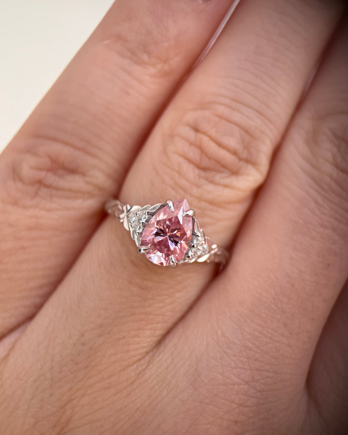 A close-up of a hand wearing a silver ring with a large, cushion-cut pink gemstone and smaller accent stones on the band-pink moissanite ring-pear engagement ring-white gold engagement ring