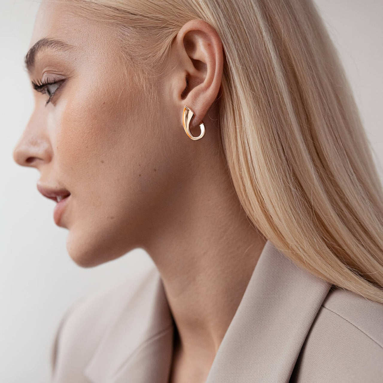 A close-up side profile of a blonde woman wearing a small gold huggie hoop earring, paired with a beige blazer against a light gray background.